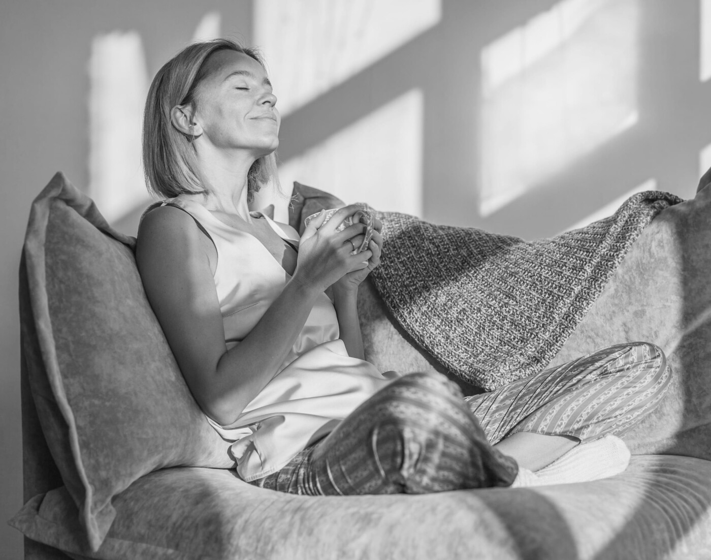 Eine Frau mit schulterlangen Haaren sitzt auf dem Sofa im Schneidersitz und hält entspannt eine Tasse in der Hand