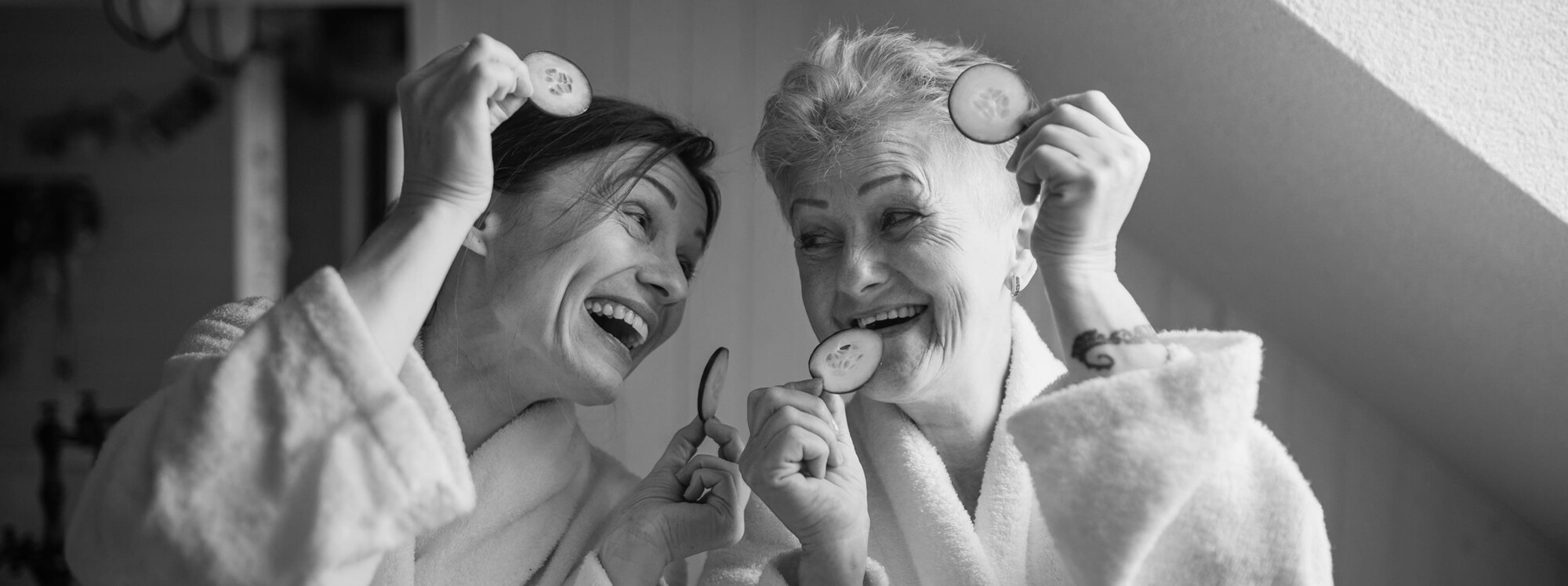 Zwei ältere Frauen im Bademantel lachen sich an und halten Gurkenscheiben in der Hand