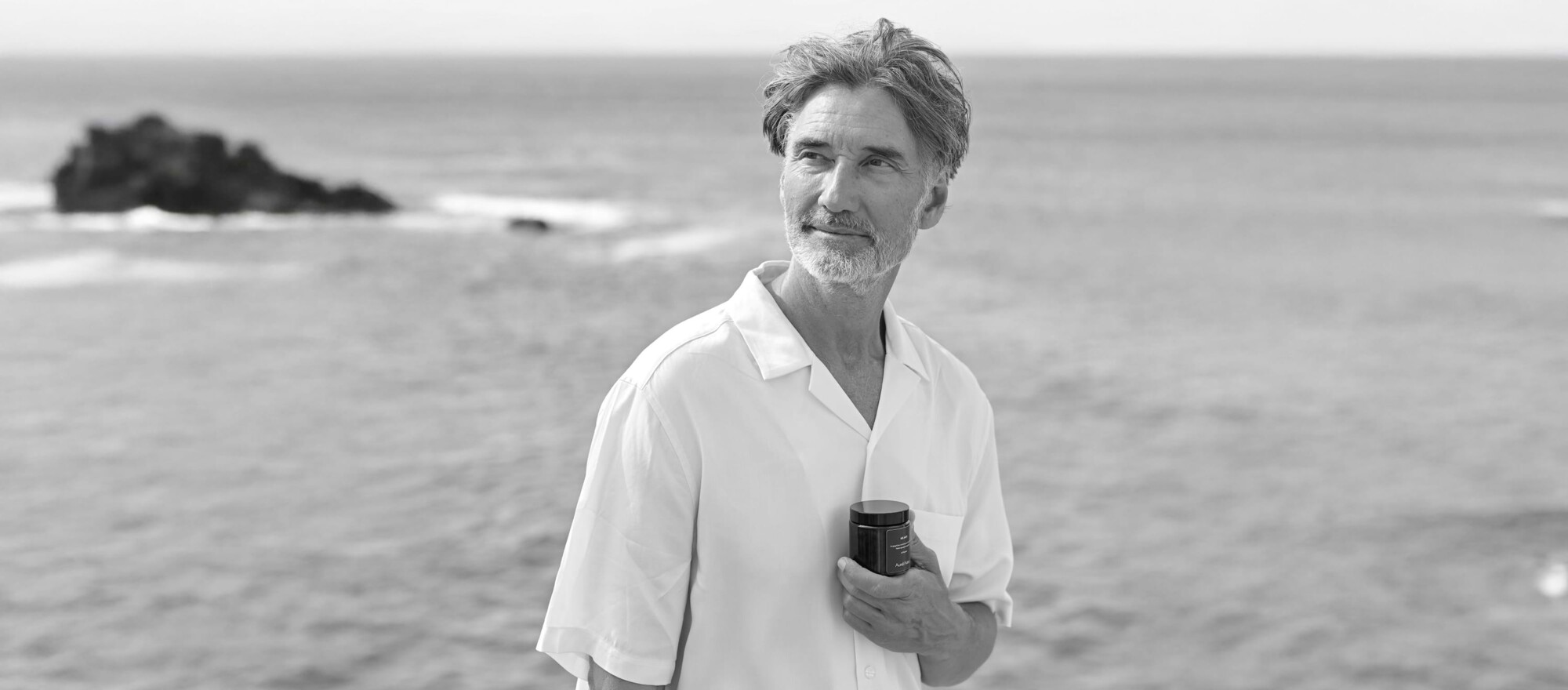 Ein älterer, gepflegter Mann steht an einer Klippe vor dem Meer und hält eine Aureli Nutra Dose in der Hand und schaut in die Ferne